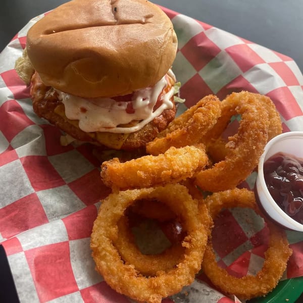 Go Vegan GR's vegan chicken sandwich with white sauce, lettuce, and tomato, served with onion rings and dipping sauce.